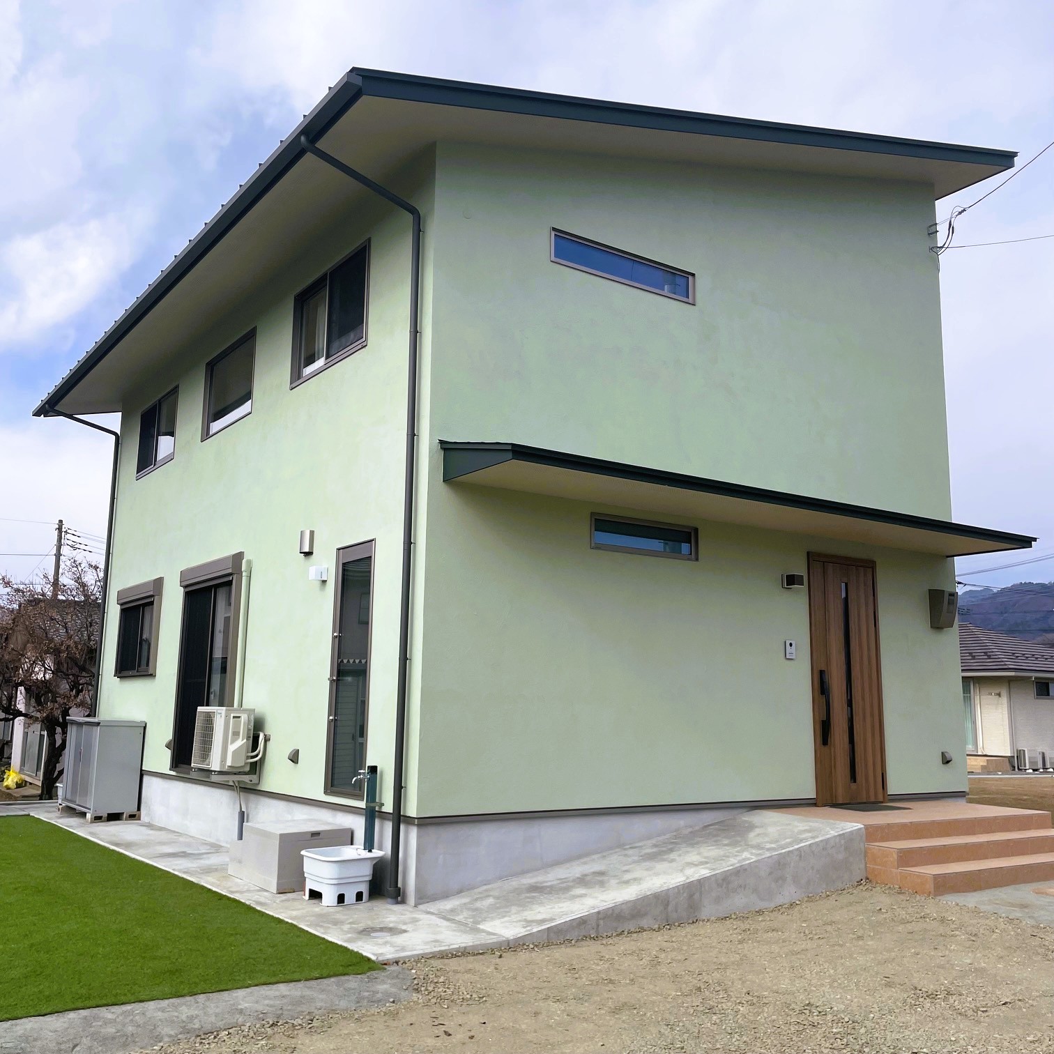 淡いグリーンの塗り壁と木製玄関ドアを組み合わせた二階建て住宅の外観(斜めアングル)