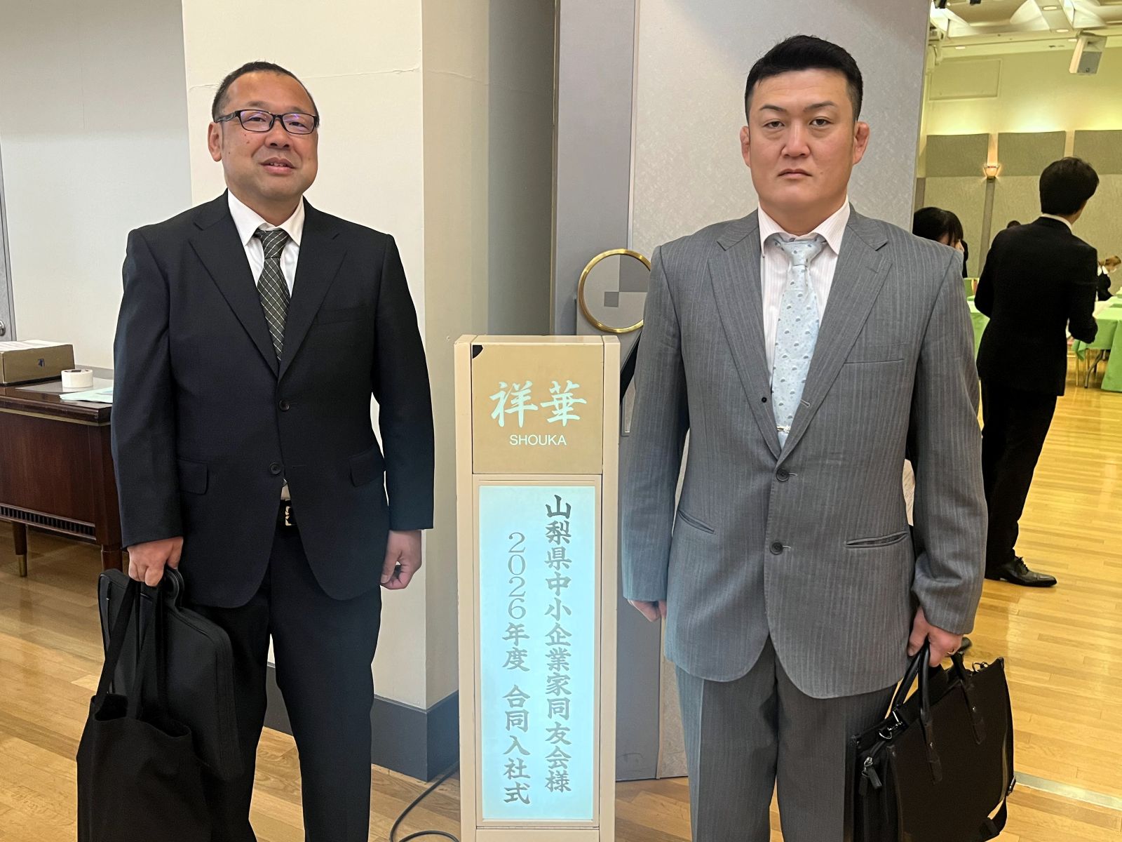 山梨県中小企業家同友会の2026年度合同入社式会場で記念撮影する山本建築工業の山本大介さんと米山亨くん