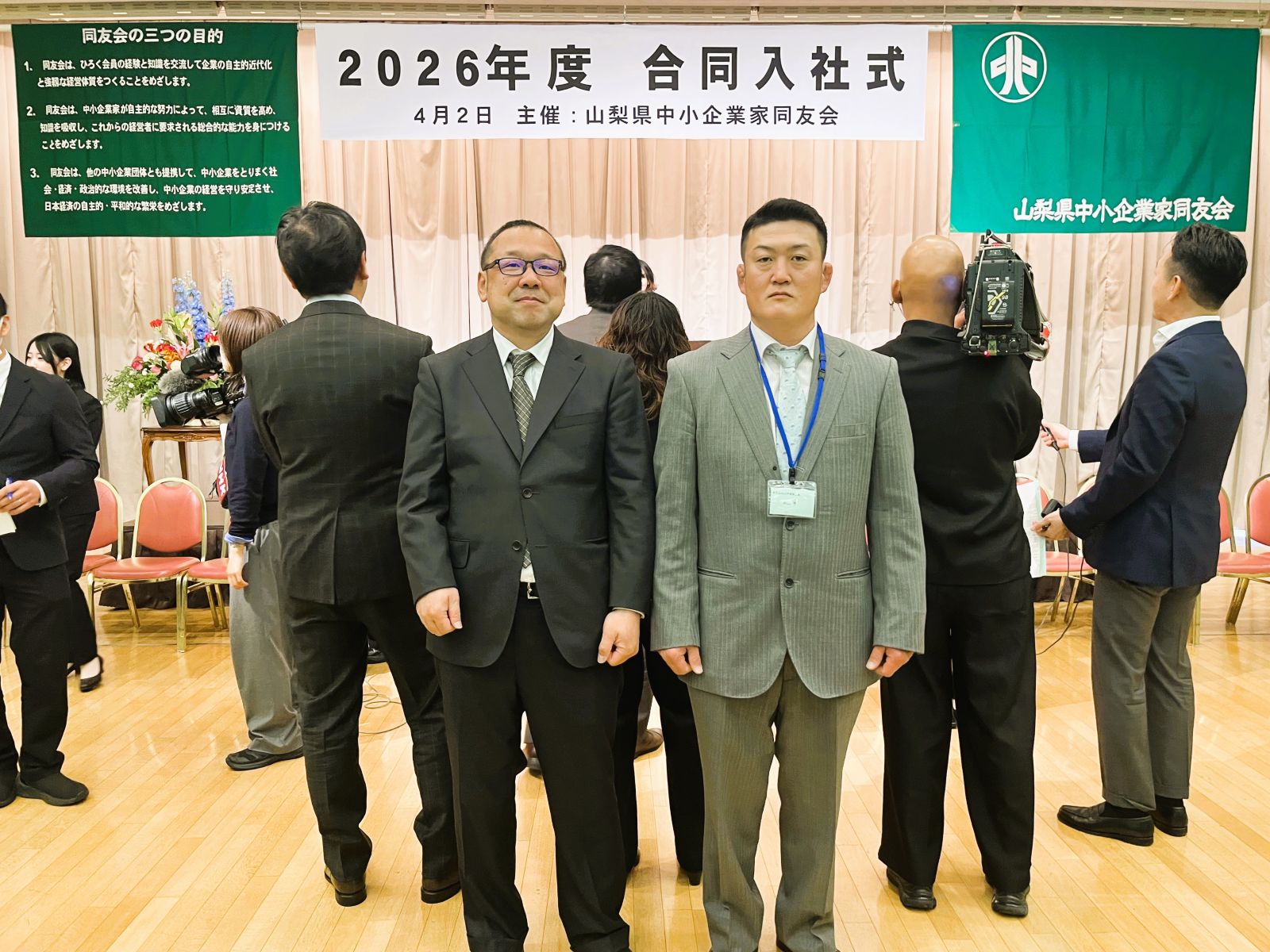 山梨県中小企業家同友会の2026年度合同入社式会場正面で撮影した山本建築工業の山本大介さんと米山亨くん