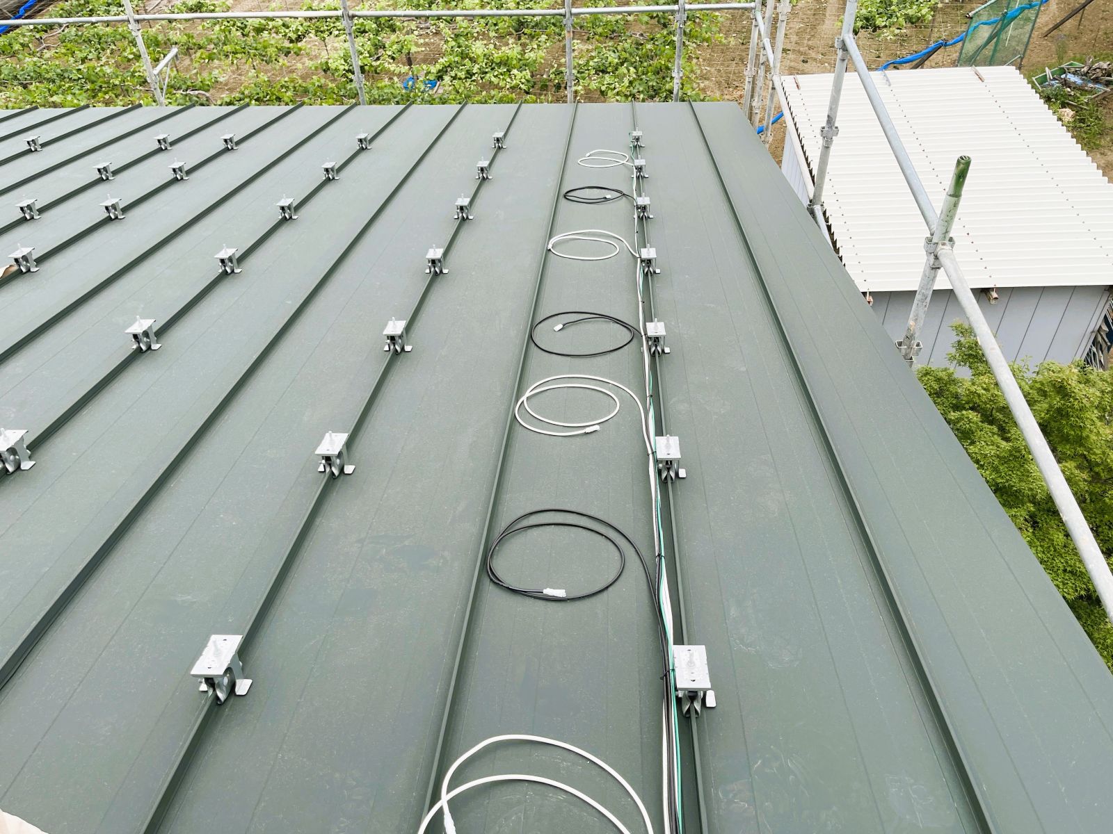 立平葺き屋根で太陽光発電設備の配線準備と支持金具取付を進めている様子