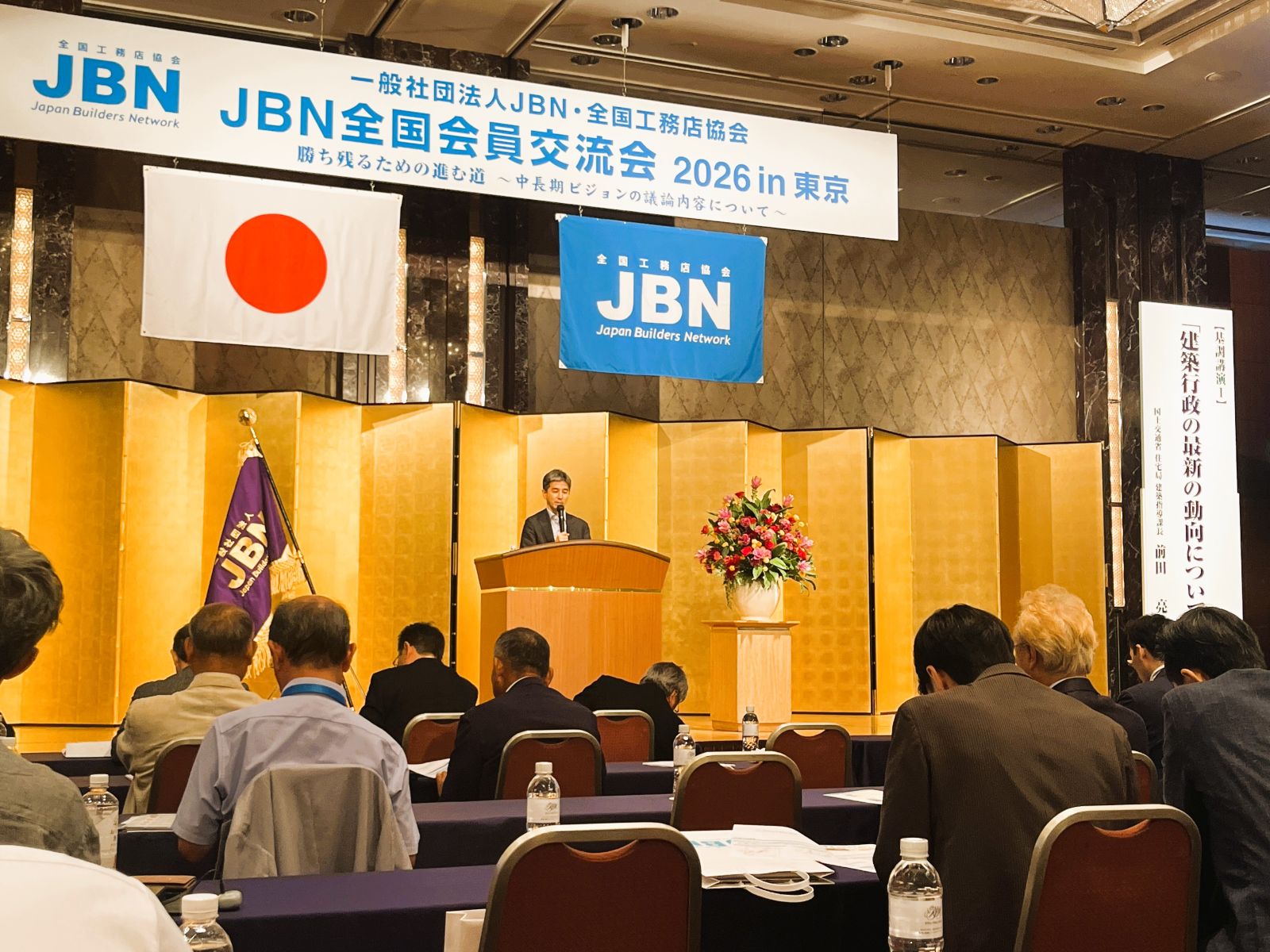 JBN全国会員交流会の会場で建築行政に関する講演を聴く参加者と演壇
